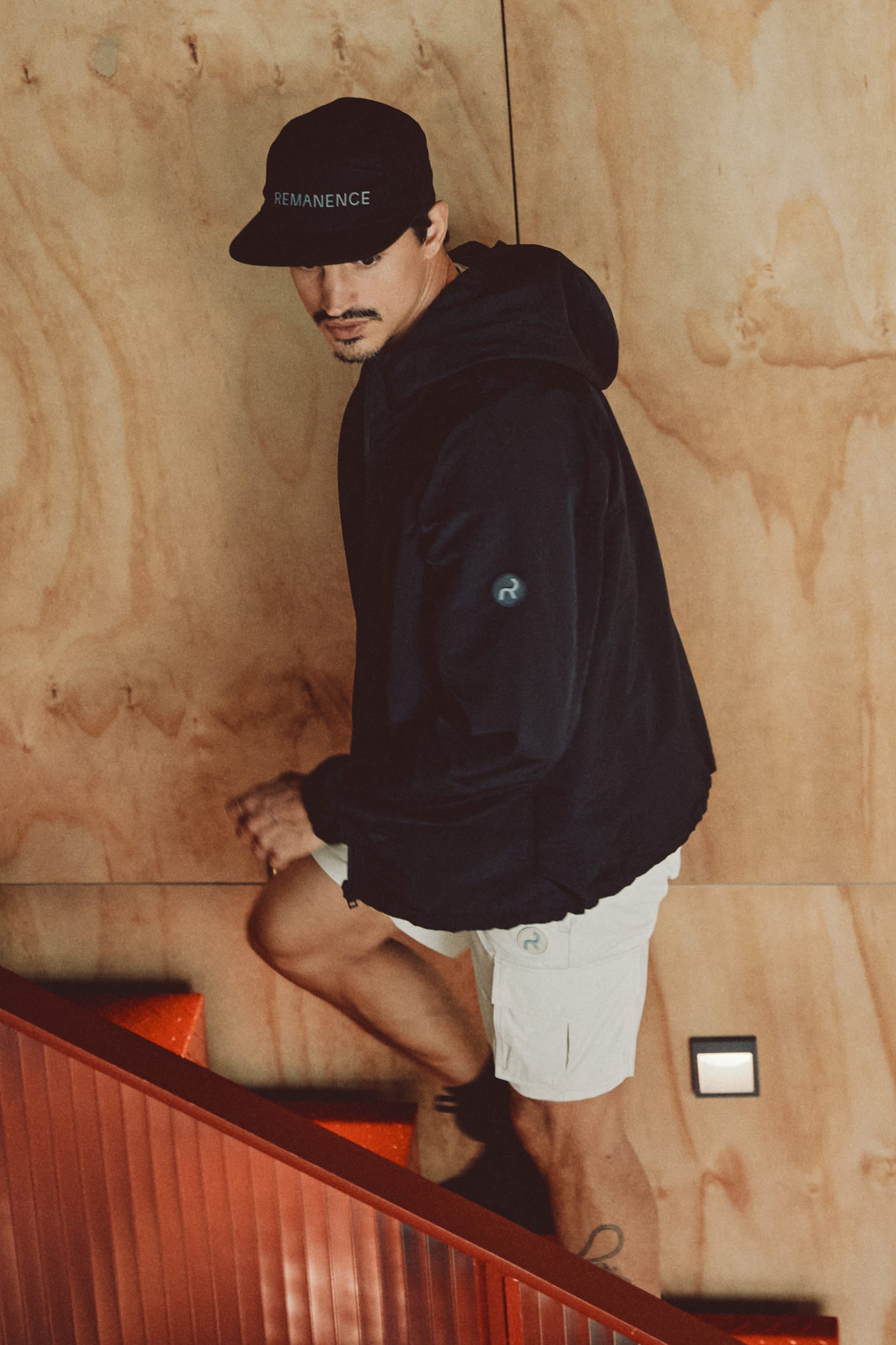 Hombre con chaqueta negra para actividades outdoor, con gorra y shorts arena remanence