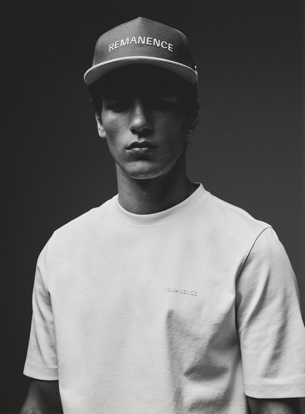 Modelo con gorra y camiseta blanca Remanence, en retrato monocromático con fondo gris oscuro.